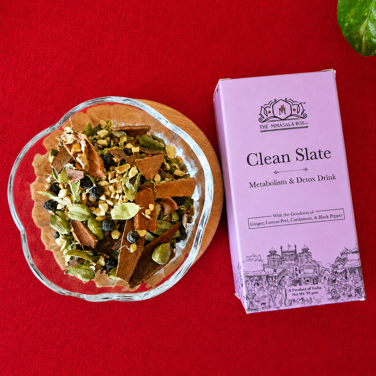 Top view of The Mmasala Box Co. Clean Slate metabolism and detox drink box placed beside a glass bowl of herbal spice blend with ginger, lemon peel, cardamom, and black pepper on a red textured background.