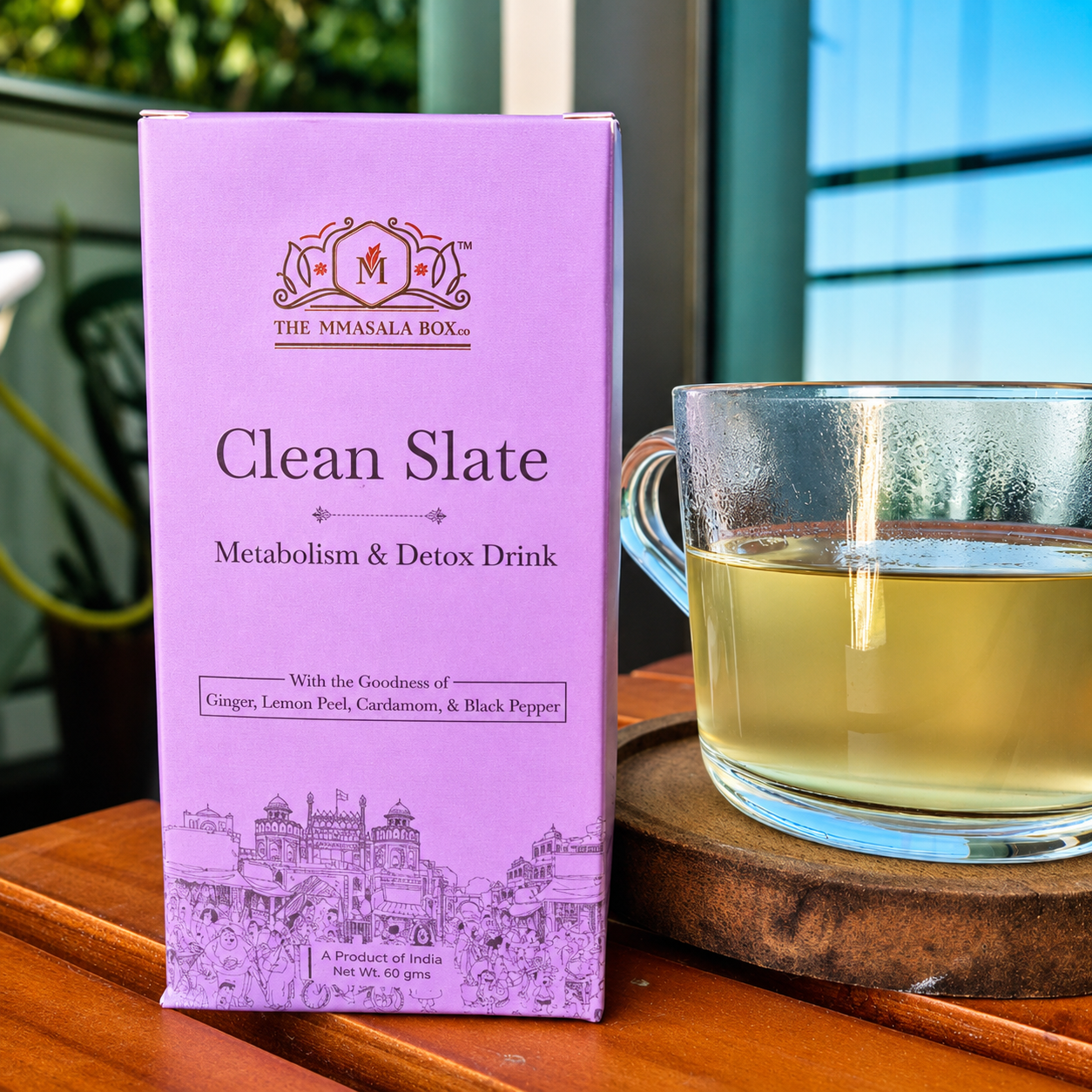 The Mmasala Box Co. Clean Slate metabolism and detox drink purple box placed beside a glass cup of herbal tea on a wooden surface, featuring ginger, lemon peel, cardamom, and black pepper ingredients.