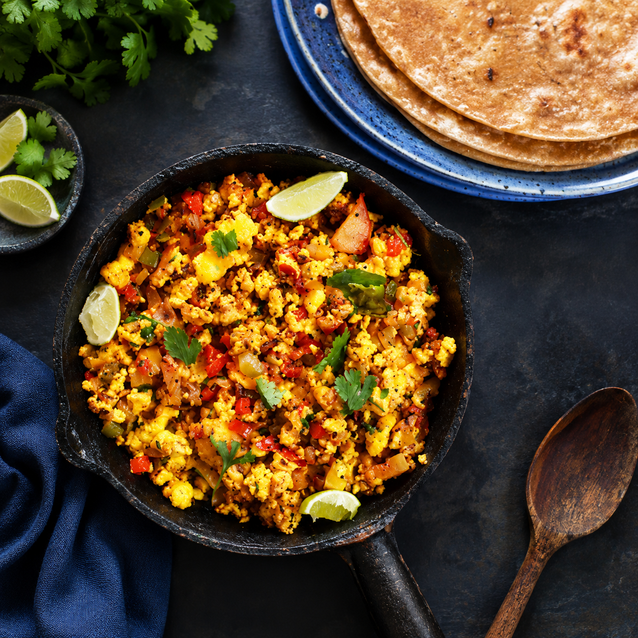 Top-view food photography of spicy paneer bhurji served in a rustic black skillet with fresh coriander and lemon wedges, paired with soft rotis on a blue ceramic plate over a dark aesthetic background.
